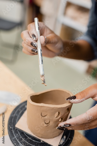 Cropped view of african american craftswoman holding tool near clay product in workshop.