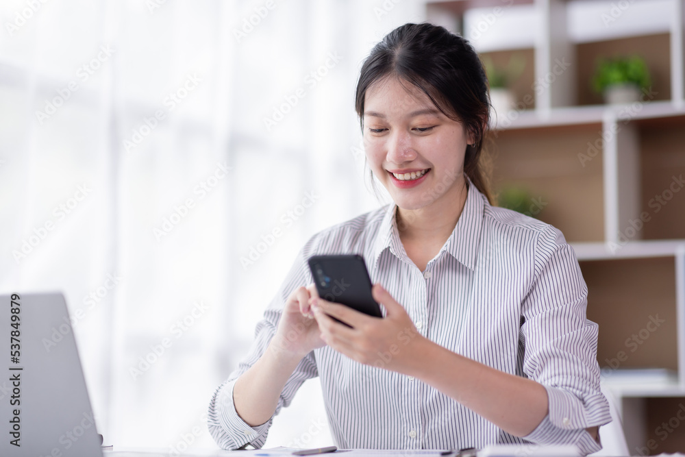 Image of smiling attractive and charming asian woman using mobile phone while working on laptop in modern office Image of smiling attractive and charming asian woman using mobile phone while working on laptop in modern office