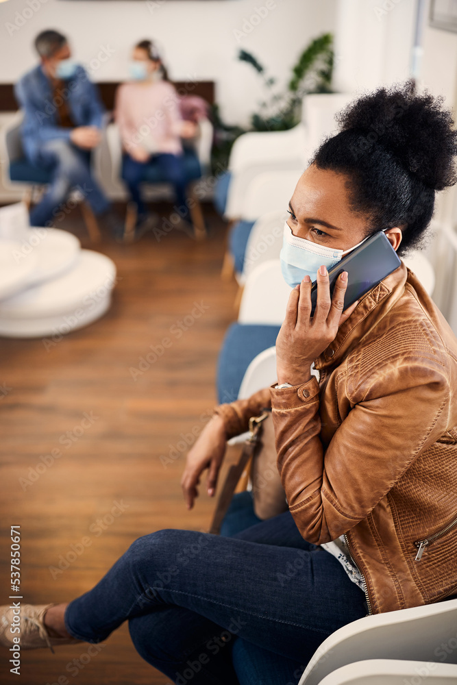 African American woman with face mask communicating over cell phone waiting for medical exam at doctor's office. African American woman with face mask communicating over cell phone waiting for medical exam at doctor's office.