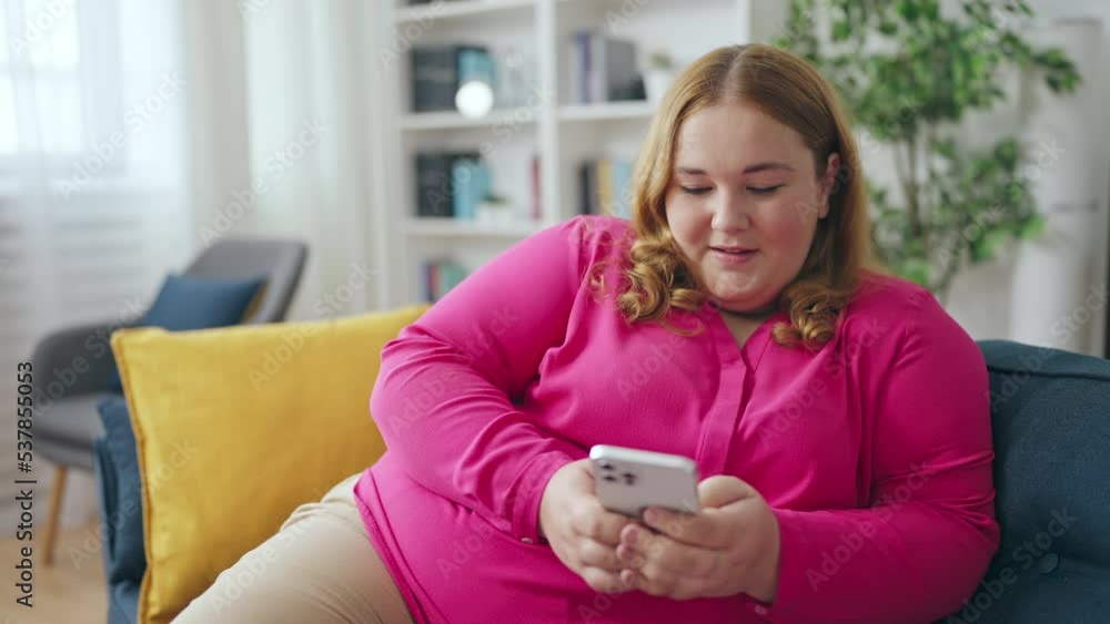 Happy plus size female using smartphone on couch, making online order, smiling Happy plus size female using smartphone on couch, making online order, smiling