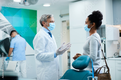 Happy dentist and black woman wear face masks while talking at dental clinic.