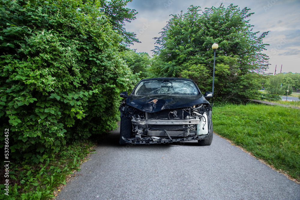 Frontal view of a black crashed car wreck - dented bonnet, smashed engine and broken windshield - in a narrow countryside summer forest road with copy space on the trees
