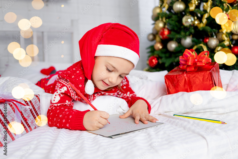 cute beautiful girl child writes a letter to Santa Claus at the Christmas tree in a red sweater and hat on New Year's Eve or Christmas at home on a white bed cute beautiful girl child writes a letter to Santa Claus at the Christmas tree in a red sweater and hat on New Year's Eve or Christmas at home on a white bed
