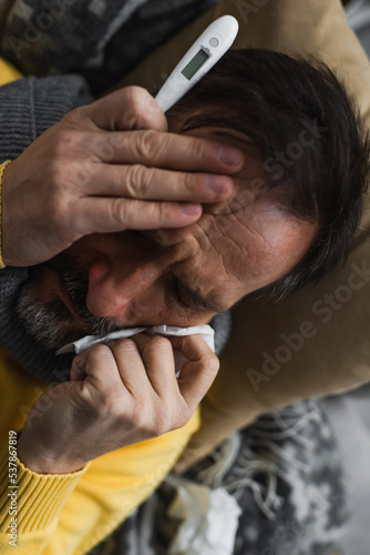 top view of diseased man with closed eyes holding paper napkin and electronic thermometer.