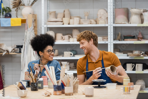 Smiling man looking at african american girlfriend near clay and paintbrushes in pottery studio. photo