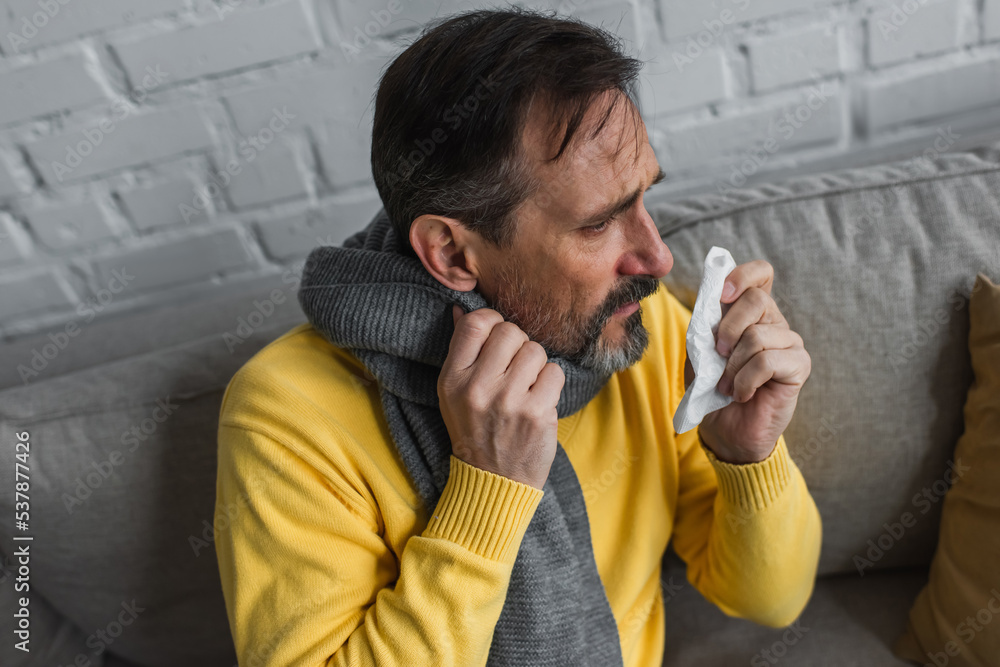 ill man in warm scarf holding paper napkin while sitting on couch at home. ill man in warm scarf holding paper napkin while sitting on couch at home.