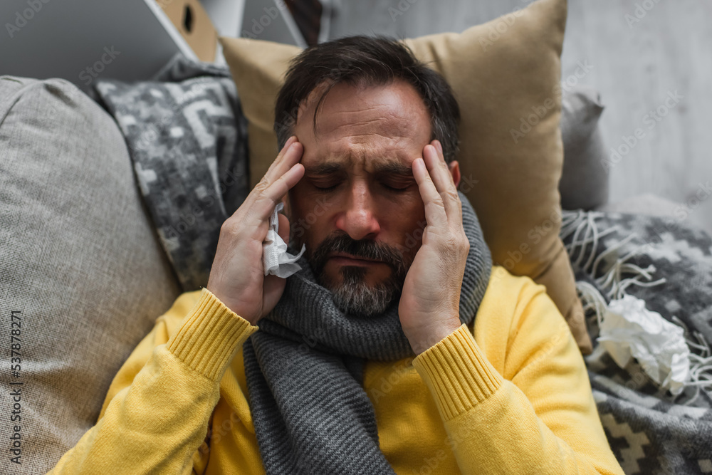 Obraz premium top view of sick man in warm scarf touching head while lying on couch and suffering from migraine.