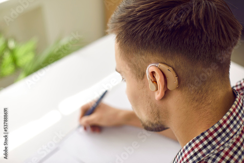 Student with hearing aid doing homework. Young deaf man wearing plastic hearing device behind his ear sitting at desk and writing. Close up of man's head. Hearing loss, impairment, education concept photo