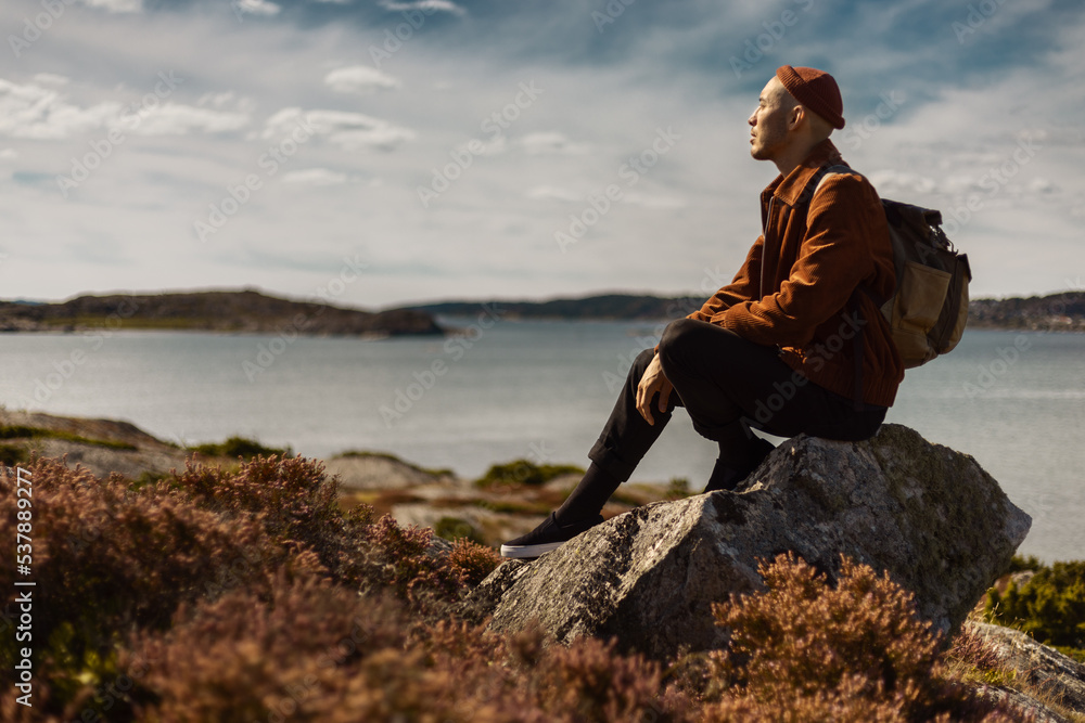 A caucasian man wearing a beanie and a brown jacket with a backpack sitting on a rock watching the sea by the coast.