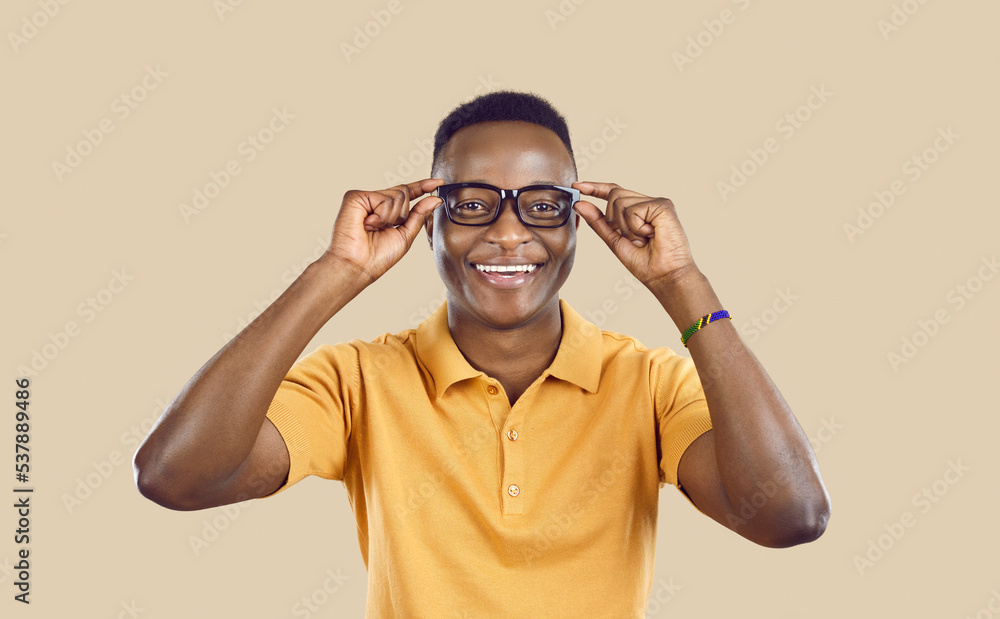 Studio shot of happy cheerful joyful handsome young African man in yellow T shirt standing on beige color background, putting on his glasses, looking at camera and smiling Studio shot of happy cheerful joyful handsome young African man in yellow T shirt standing on beige color background, putting on his glasses, looking at camera and smiling