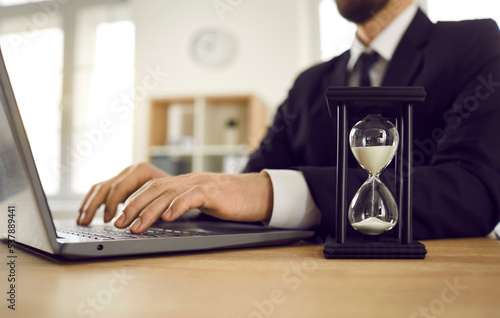 Man working on notebook computer at desk with hour glass. Employee typing on PC keyboard and trying to meet deadline. Don't waste time, it's a limited resource. Carpe diem. Seize the day. Seize moment photo