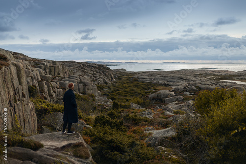 A man wearing a blue coat and a brown beanie standing on a cliff by the coast watching a rocky landscape and the sea.