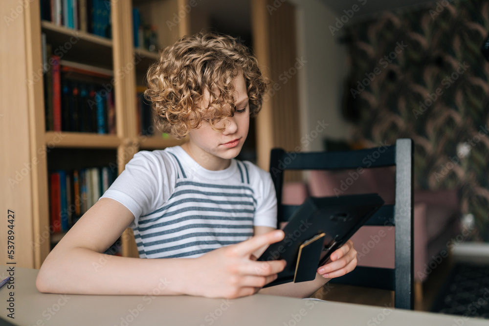 Close-up of upset preteen girl holding photo frame in hands and looking sorrowfully at family portrait. Frustrated adorable kid missed person in picture. Concept of nostalgia, grief, longing and loss Close-up of upset preteen girl holding photo frame in hands and looking sorrowfully at family portrait. Frustrated adorable kid missed person in picture. Concept of nostalgia, grief, longing and loss