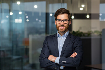 Portrait of successful mature boss, senior businessman in glasses and business suit looking at camera and smiling, man with crossed arms working inside modern office building.
