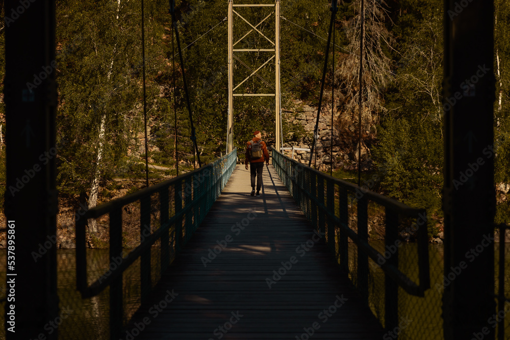 Fototapeta premium Man with backpack walking across a narrow suspension bridge over a river in a lush forest during golden hour. Cinematic view of outdoor adventure, hiking, and exploration in nature.