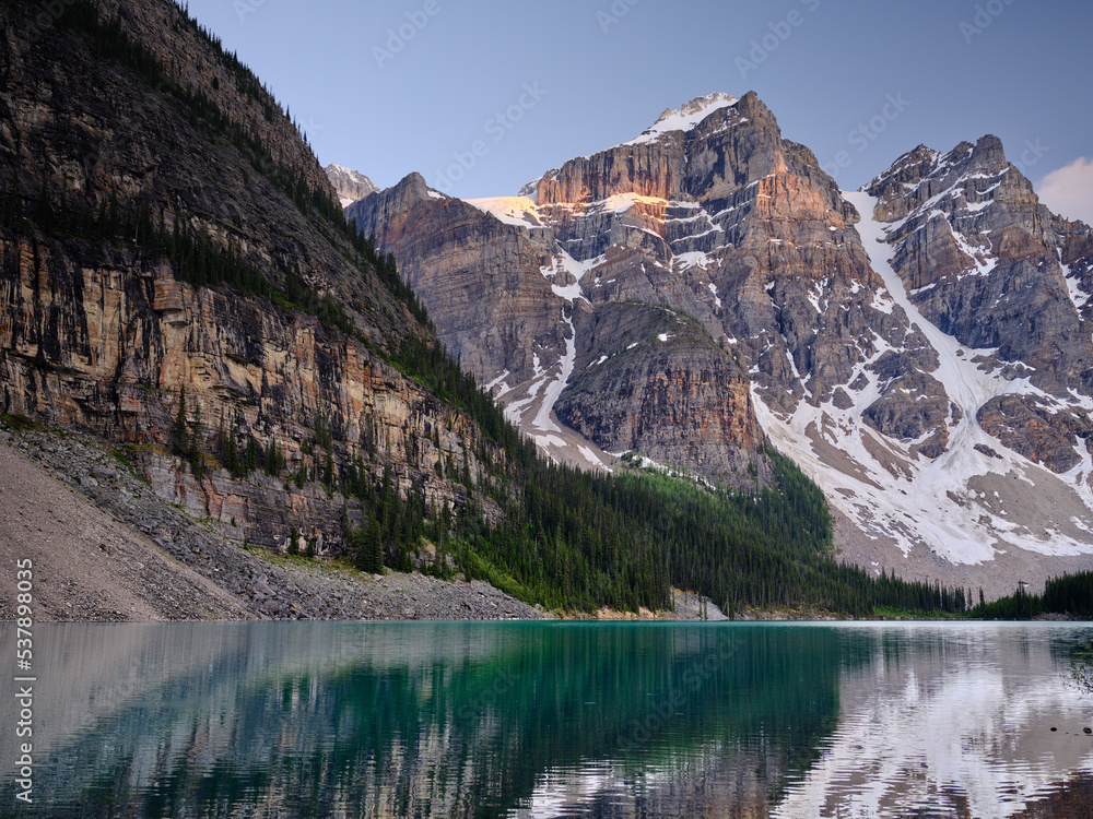 Sunset lights up the mountain peaks of the Three Sisters on the clear turquoise waters of Moraine lake Canada