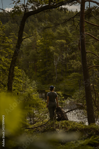 A woman wearing a hat standing and a back pack at a lookout point in the forest watching over a lake in summer.