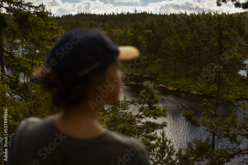 Fototapeta Naklejka Na Ścianę i Meble -  A woman wearing a hat standing at a lookout point in the forest watching over a lake in summer.