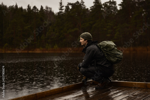 Quadro su tela A hiker in rain gear and a beanie squats on a wooden pier by a misty lake in a dark forest