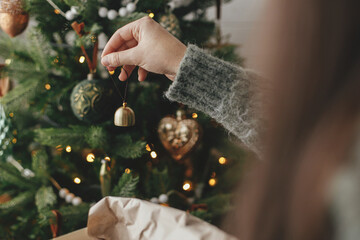 Hands decorating christmas tree with stylish bauble in atmospheric festive room. Merry Christmas! Winter holidays preparation. Woman in cozy sweater putting vintage bell on tree