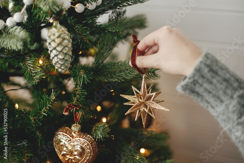 Hands decorating christmas tree with stylish bauble in atmospheric festive room. Merry Christmas! Winter holidays preparation. Woman in cozy sweater putting vintage star on tree