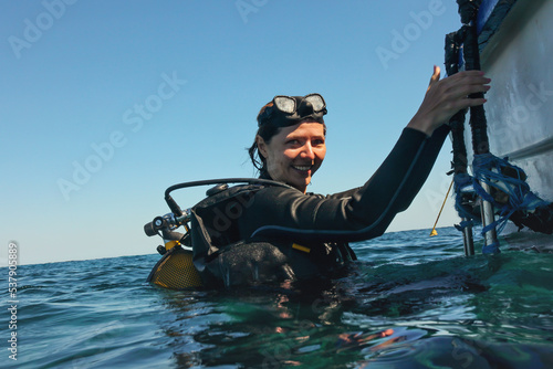 Young woman in neoprene, smiling, mask on head, getting to boat from sea after her dive