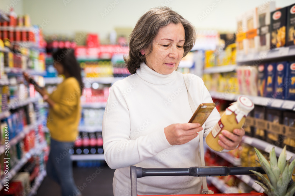 Positive interested elderly woman scanning barcode on bottle of fruit juice with her smartphone, checking product information. Concept of modern technologies for convenience of buyers