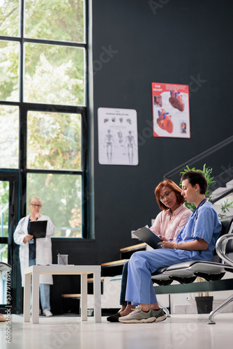 Medical asisstant consulting senior patient in hospital waiting area, filling report papers explaining healthcare treatment. Nurse doing checkup examination with asian woman in lobby. photo