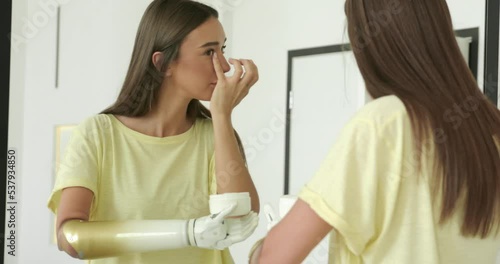 Wallpaper Mural woman with prosthetic arm applying cream near mirror at home, girl with disability using artificial prosthetic limb to hold cream Torontodigital.ca
