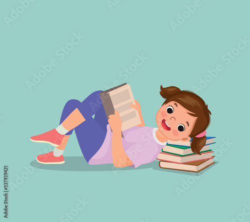 Cute little girl reading book lying on the floor with head on stack of books 