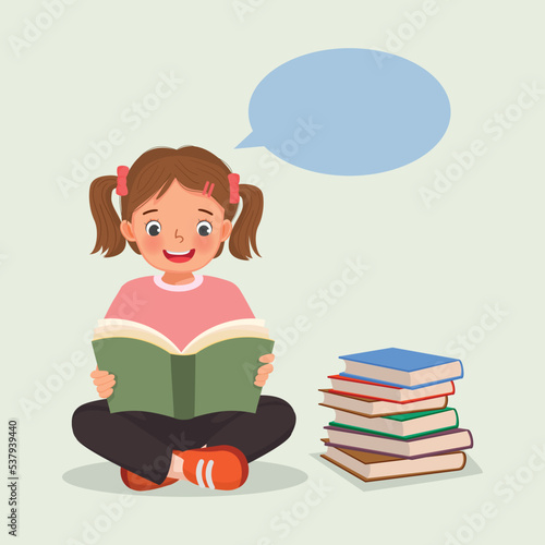 Cute little girl reading a book sitting on the floor beside stack of books