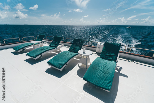 A row of four deckchairs with teal cushions on them and two small coffee tables in between on the upper deck of a luxurious diving safari boat or a recreational vessel lit by bright Maldivian sun photo
