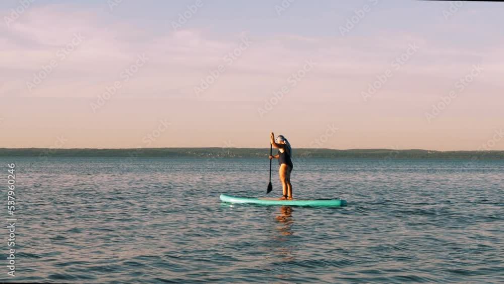A woman in a swimsuit and a turban floats on a SUP board on smooth water on a clear day. Overall plan A woman in a swimsuit and a turban floats on a SUP board on smooth water on a clear day. Overall plan