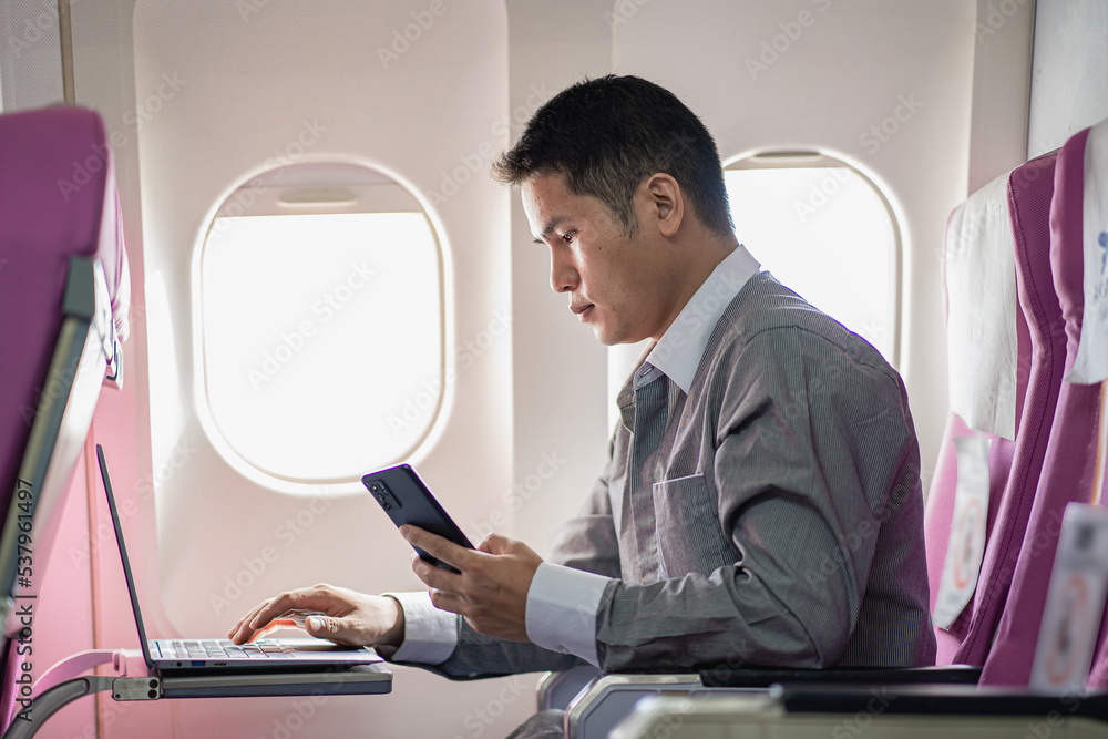 Fototapeta premium young man working on plane and holding smartphone and laptop while sitting in the cabin of an airplane work and travel concept