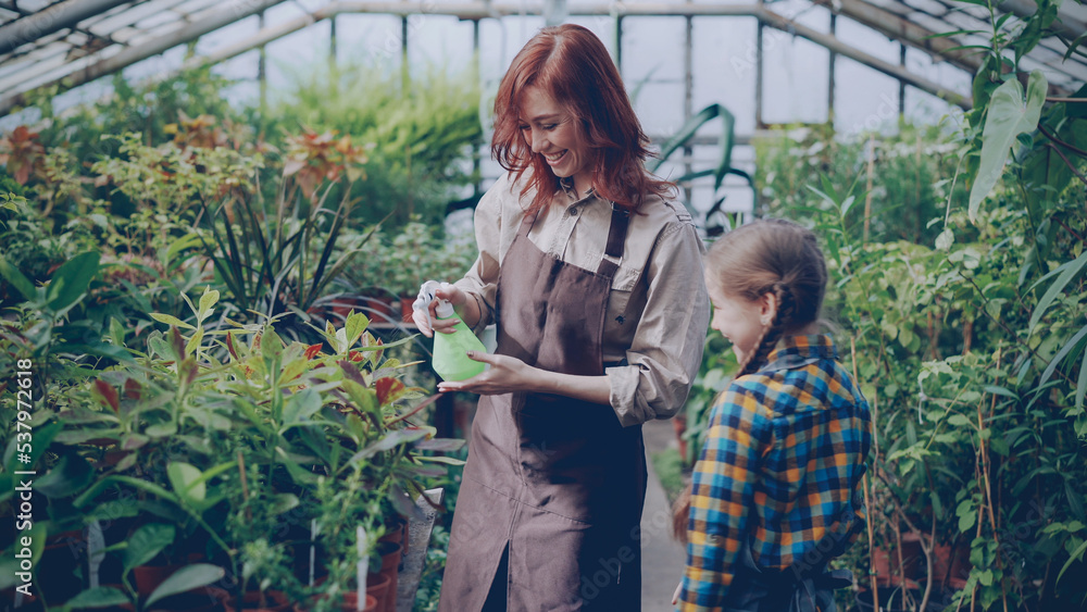 Young woman and her funny daughter are sprinkling water on flowers, child is having fun and spraying her mother with laughter. Growing plants and happy family concept.