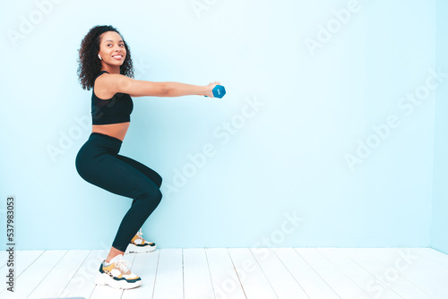 Fitness smiling black woman in sports clothing with afro curls hairstyle. She wearing sportswear. Young beautiful model doing squats. Female holding dumbbells in studio near light blue wall