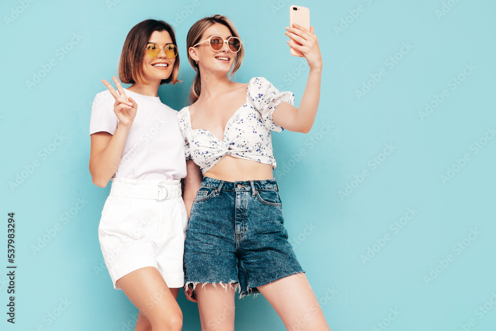 Two young beautiful smiling brunette hipster female in trendy summer shorts clothes. Sexy carefree women posing near blue wall. Positive models having fun. Cheerful and happy. Hugging. Takes selfie Two young beautiful smiling brunette hipster female in trendy summer shorts clothes. Sexy carefree women posing near blue wall. Positive models having fun. Cheerful and happy. Hugging. Takes selfie