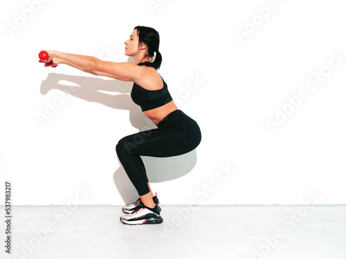 Fitness smiling woman in black sports clothing. Sexy young beautiful model with perfect body. Female isolated on white wall in studio. Stretching out before training.Making squats with dumbbells