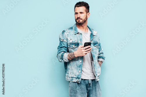 Portrait of handsome confident stylish hipster lambersexual model. Sexy man dressed in jeans jacket. Fashion male isolated on blue wall in studio. Holding smartphone