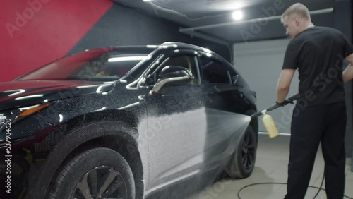 Black automobile on a car wash. Caucasian man in dark clothes using high pressure washer and snow foam cannon to spray vehicle with water and shampoo, pre-wash. Indoors, carwash service photo