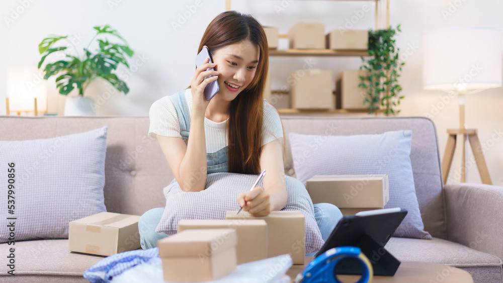 Online merchant concept, Female entrepreneur talks on smartphone and writes address on parcel boxes Online merchant concept, Female entrepreneur talks on smartphone and writes address on parcel boxes