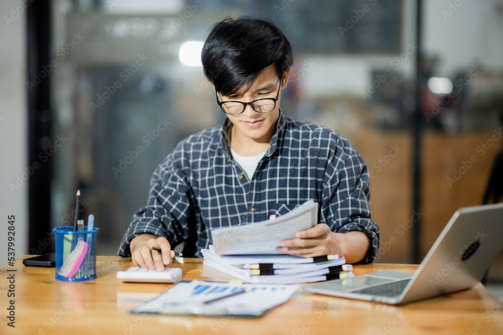A young financial market analyst works in the office with his laptop while sitting at a wooden table. businessman analyzing graph document in hand. A young financial market analyst works in the office with his laptop while sitting at a wooden table. businessman analyzing graph document in hand.