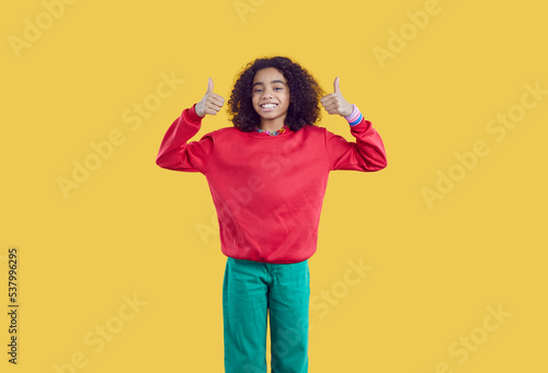 Happy african american pretten girl on yellow background showing thumbs up recommending best choice. Portrait of positive cute curly ethnic teen girl dressed in casual clothes showing two thumbs up.