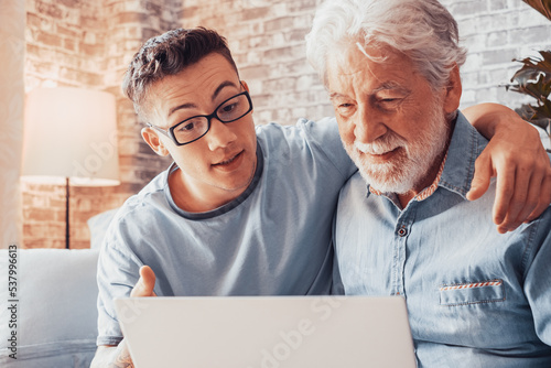 Принти на полотні Smiling young boy helping senior grandfather browse with laptop and choose goods or services on web surfing together at home