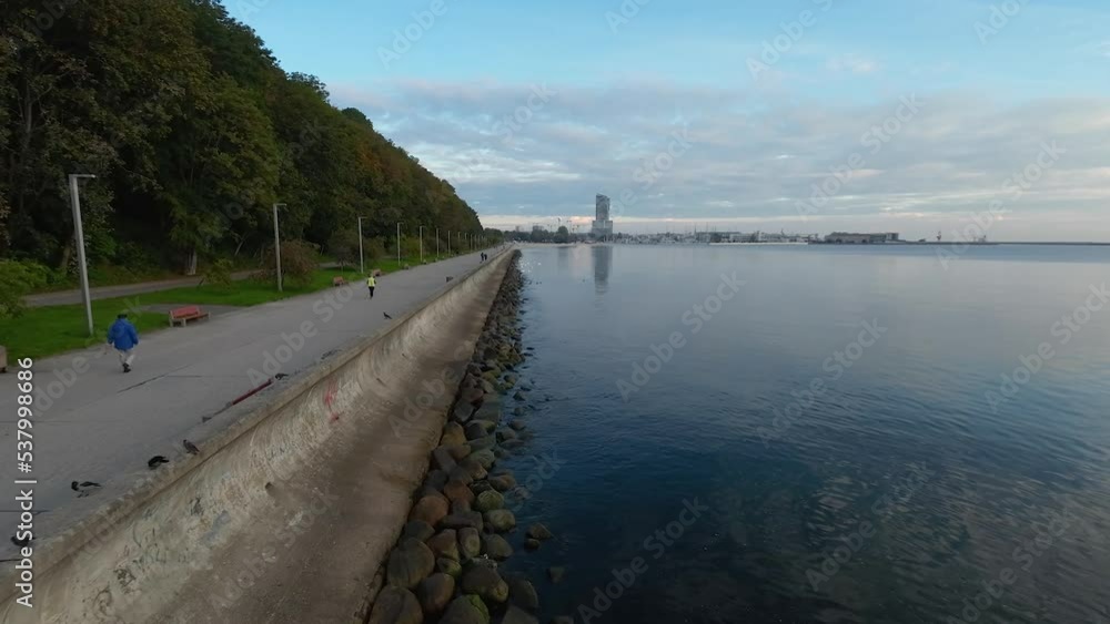 People walking on Boulevard in Gdynia, low drone flight over coast, birds start from off the coast. People walking on Boulevard in Gdynia, low drone flight over coast, birds start from off the coast.
