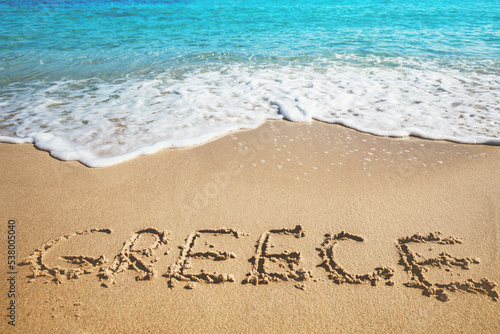 Inscription GREECE on a sandy beach with sea waves. Nature background