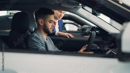 Confident man car buyer is sitting in driver's seat in new automobile inside modern showroom and talking to salesman standing near car window. Guy is holding steering wheel. © silverkblack