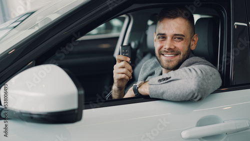 Close-up portrait of handsome bearded guy new car owner sitting inside beautiful automobile holding key fob and smiling looking at camera. Transportation and people concept.