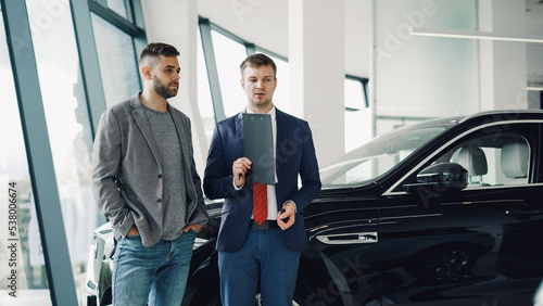 Handsome bearded guy is talking to car dealership manager in motor show discussing new automobile model, dealer is holding documents and pen. Purchasing vehicle and people concept. © silverkblack