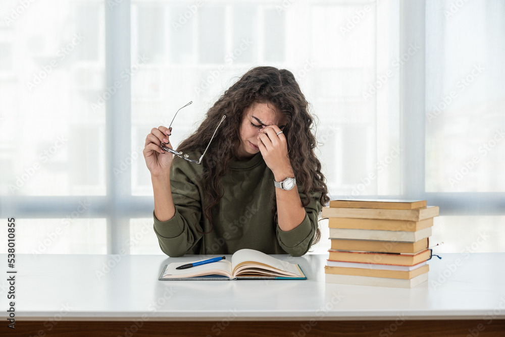 Young frustrated school girl sitting at desk with pile of books feeling sad that she need to read. Female first year college student get depressed from how much she need to study. Education concept Young frustrated school girl sitting at desk with pile of books feeling sad that she need to read. Female first year college student get depressed from how much she need to study. Education concept
